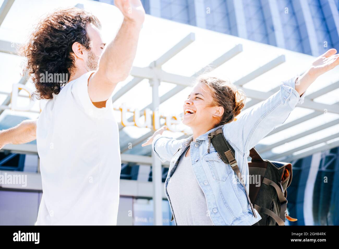 Happy interracial couple at the airport. Husband waving and raising hands in salute meeting wife after business trip and embracing at departure area Stock Photo