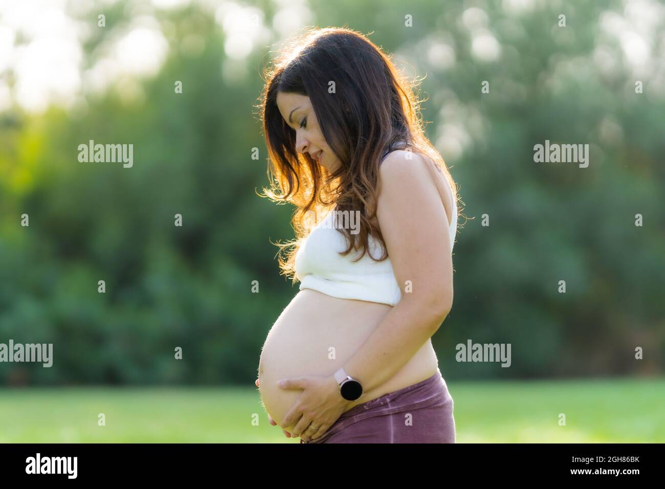 Pregnant woman touching her abdomen in a park with the sunlight shining on her Stock Photo