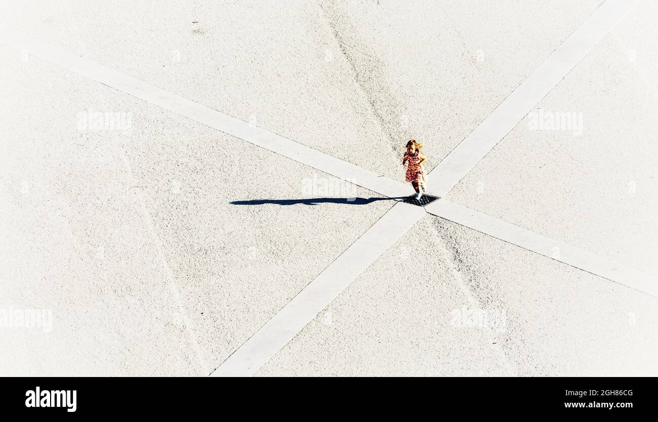 Top view of a blonde girl in flower dress running across the diagonal of a monochrome deserted square Stock Photo