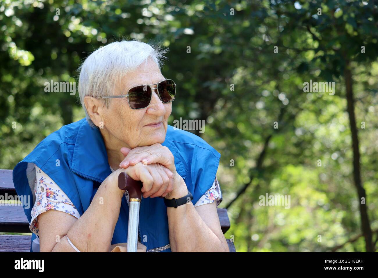 Happy old woman in sunglasses sitting with walking cane on a bench in park. Life in retirement, enjoying the nature Stock Photo