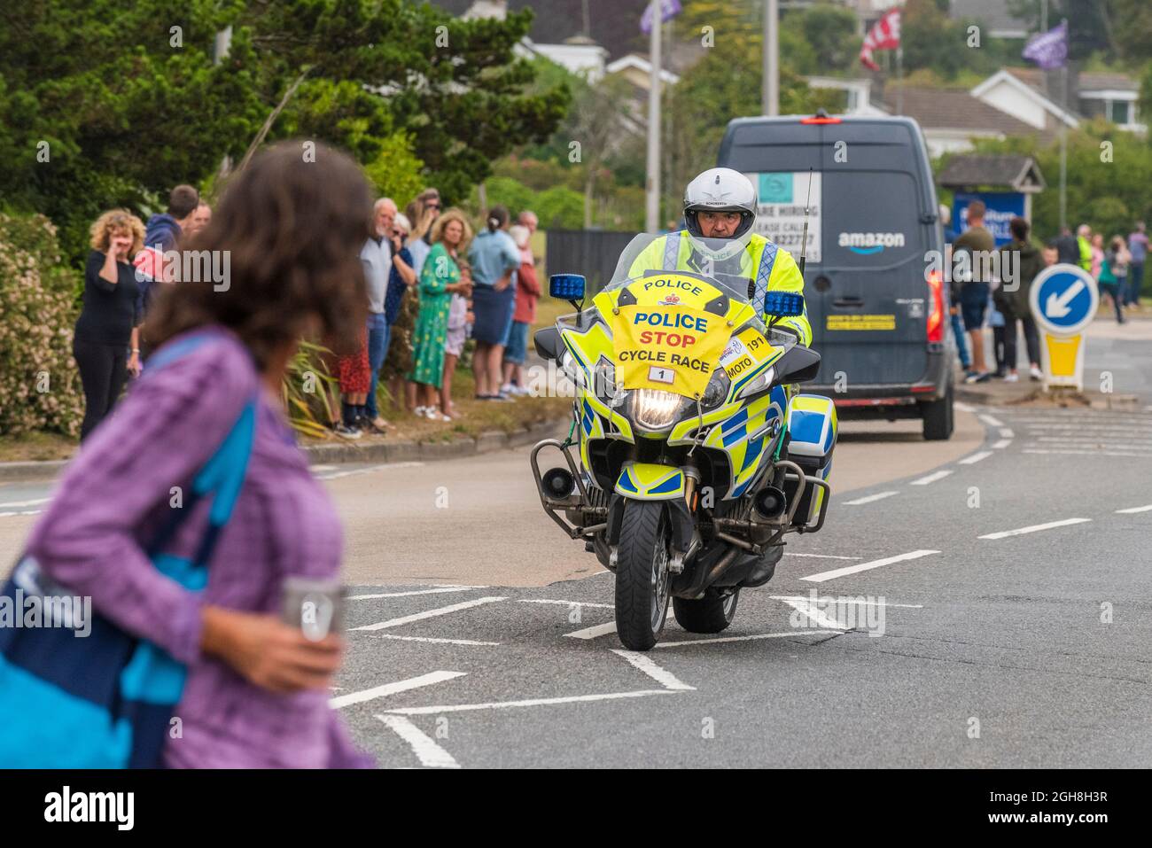 A pedestrian walking across a road in front of police patrol motorcyclist riding into Newquay in Cornwall during the opening stage of the iconic Tour Stock Photo