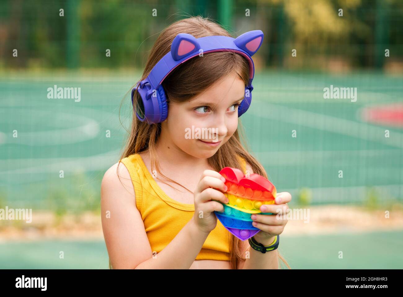 Portrait young girl in wireless headphones on sports ground playing with the Pop It fidget. Push pop bubble flexible fidget sensory toy. Feel no stress free Stock Photo