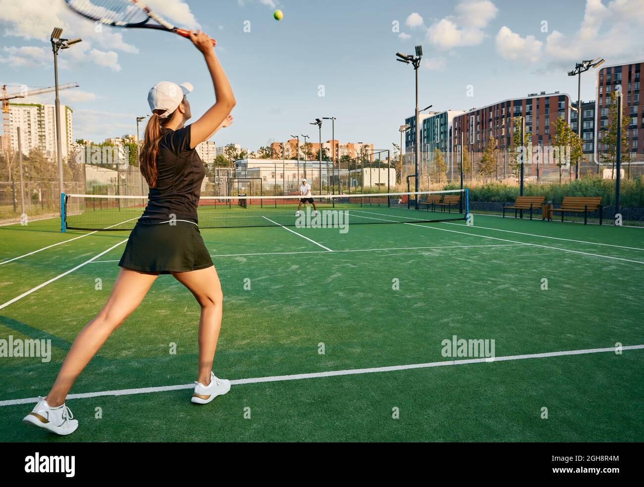 tennis player serves ball while playing match with her male partner on a grass court in urban environment. Female tennis player with tennis racket and Stock Photo