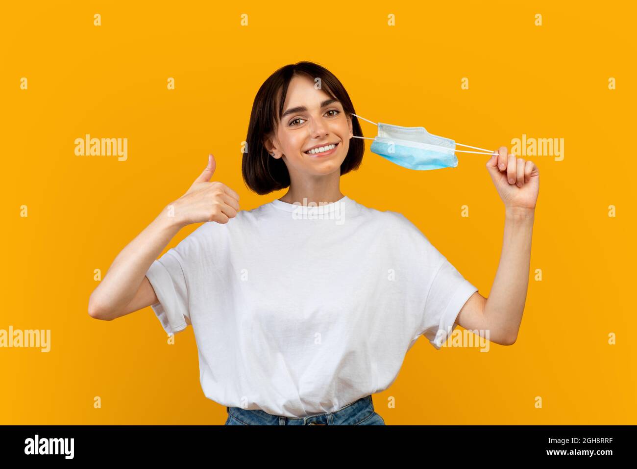 Coronavirus is over. Happy woman taking off her face mask and showing thumb up gesture on yellow studio background Stock Photo