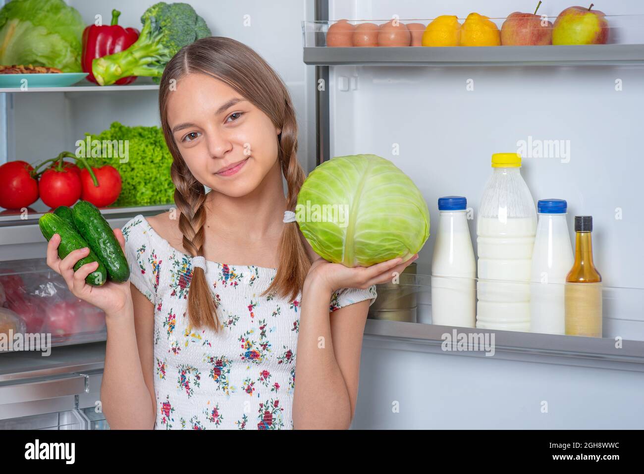 Beautiful young teen girl holding fresh green cabbage and green cucumbers while standing near open fridge in kitchen at home Stock Photo