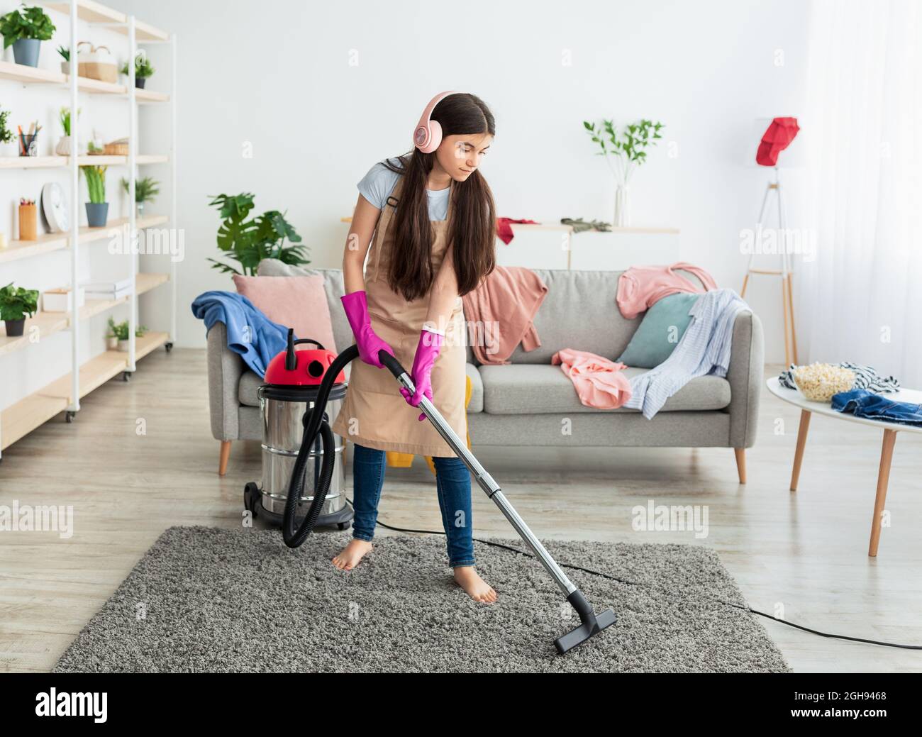 Full length of Indian teen girl cleaning apartment, wearing headphones, listening to music while vacuuming floor Stock Photo