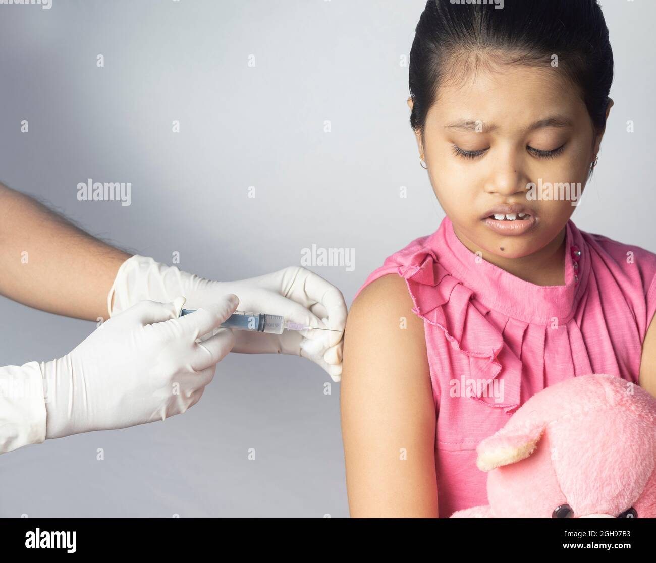 An Indian girl child with teddy bear in lap receiving vaccine dose on white background Stock Photo