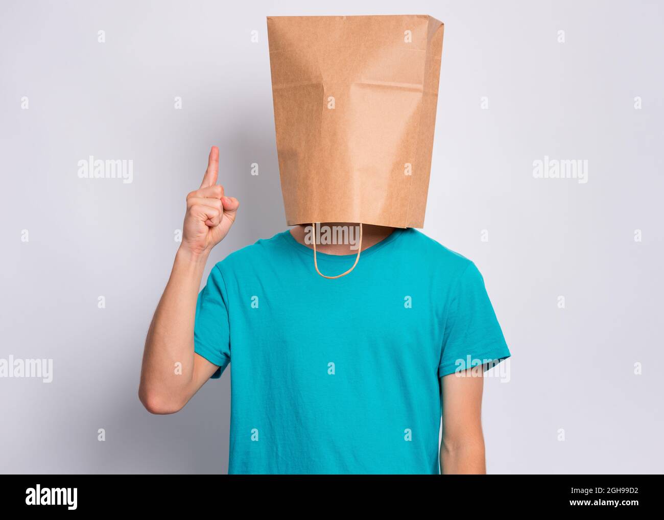 Portrait of teen boy with paper bag over head pointing hand up at copyspace, on gray background. Child pointing finger at something. Stock Photo