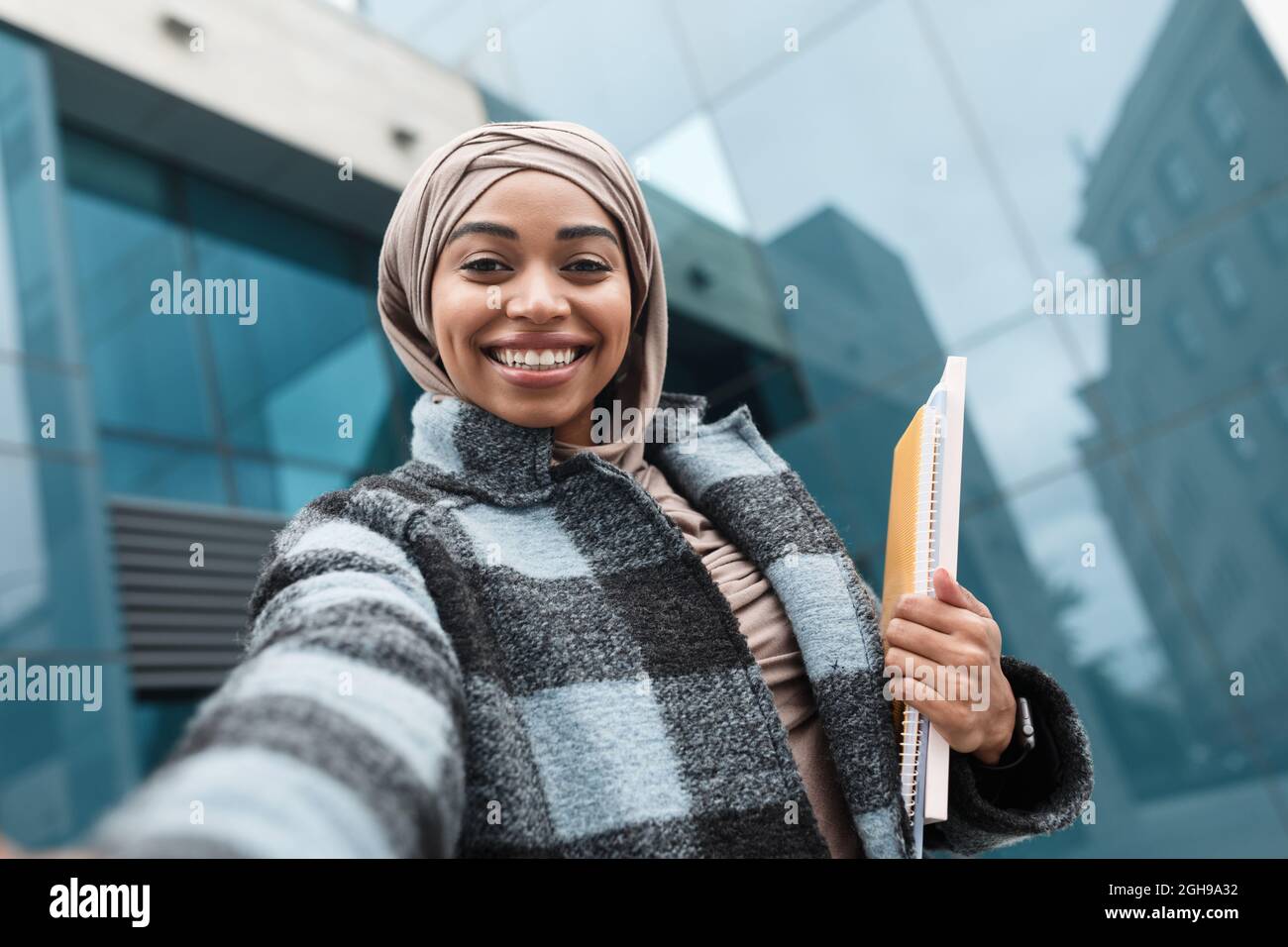 Satisfied attractive islamic afro american woman student in hijab and coat with books looks at gadget webcam Stock Photo