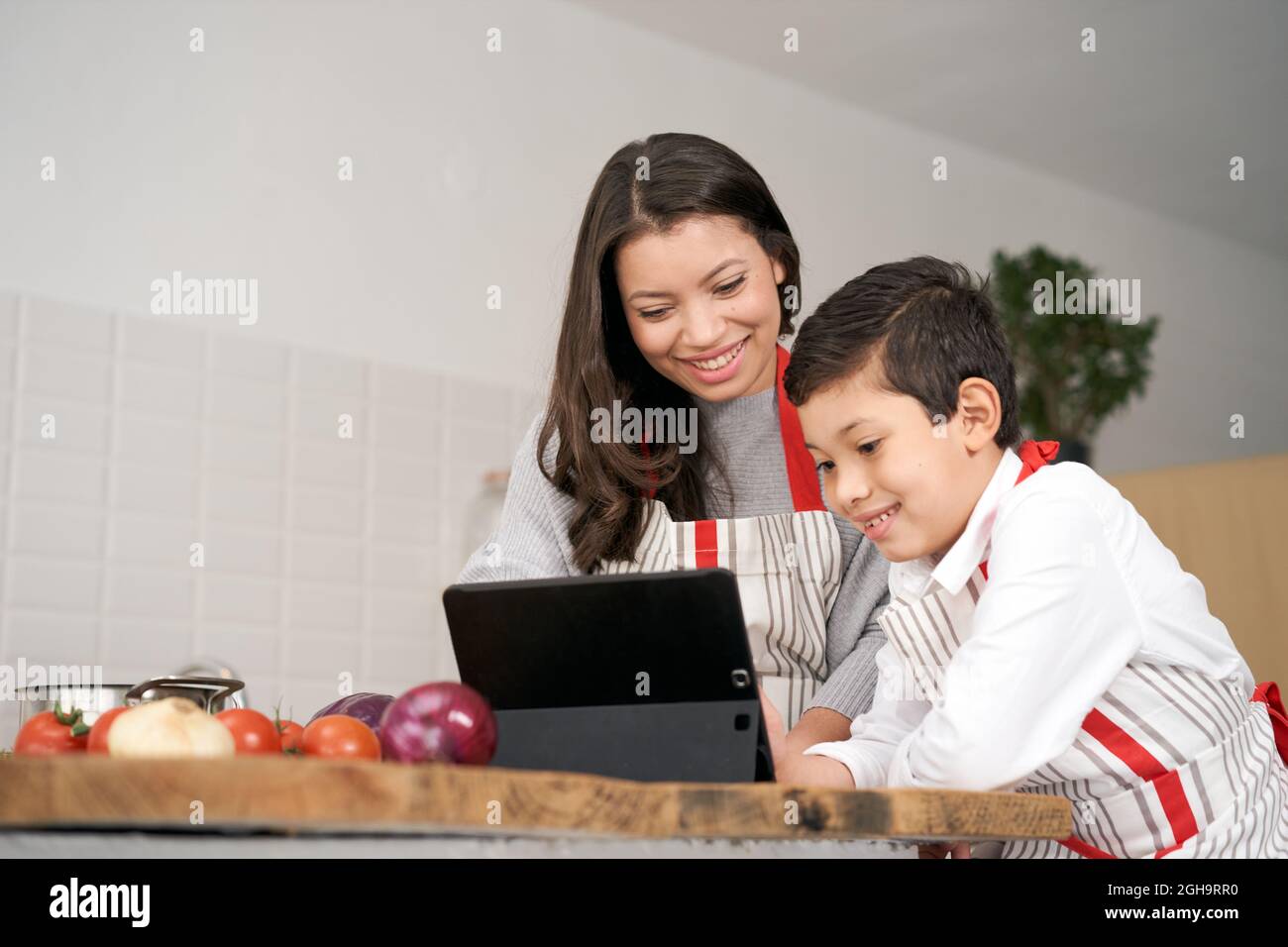Mother and son search for healthy food recipes on the internet while cooking some vegetables in the kitchen. Lifestyle with latin people. Stock Photo