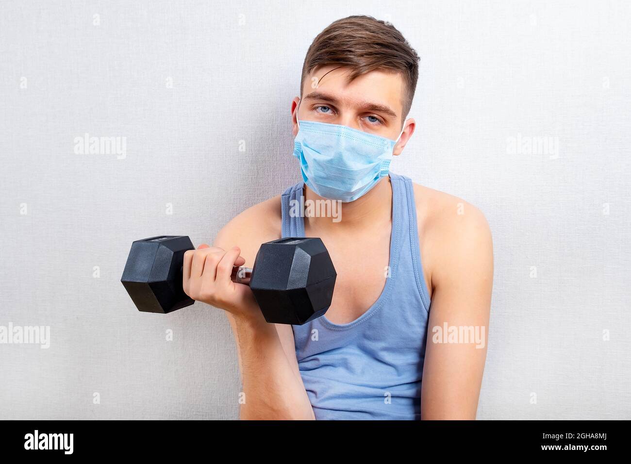 Sad and Weak Young Man in Flu Mask hold a Dumbbell by the White Wall ...