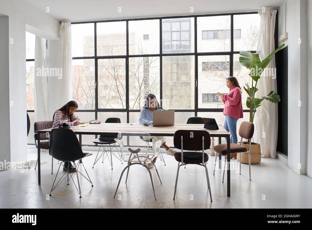 In the office. Three businesswomen work together without masks, keeping a safe distance. Two of them are working at the desk and one is chatting on Stock Photo