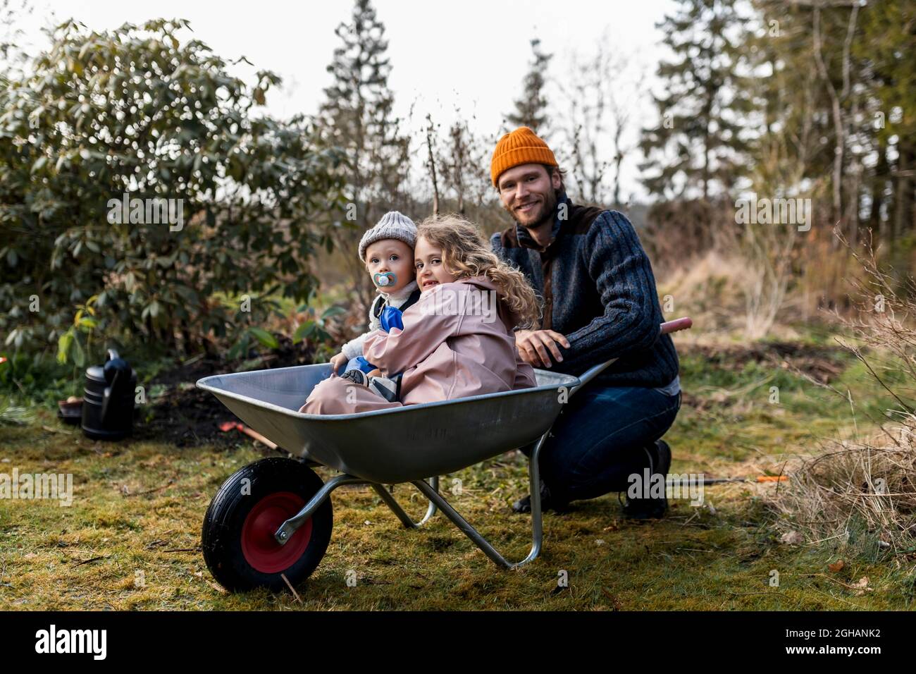 Boy and girl sitting in wheelbarrow by father at back yard Stock Photo