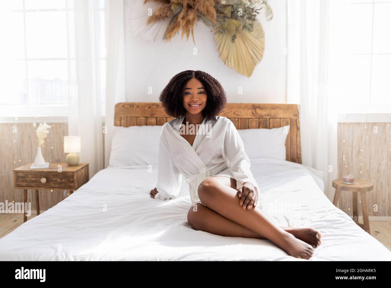 Millennial black woman in dressing gown showing legs after epilation, enjoying soft skin on bed at home Stock Photo