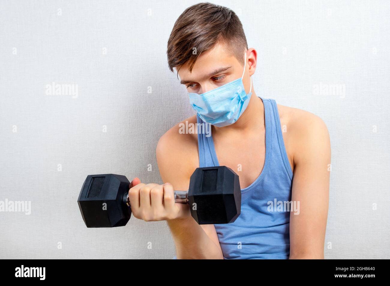 Sad and Weak Young Man in Flu Mask hold a Dumbbell by the White Wall ...