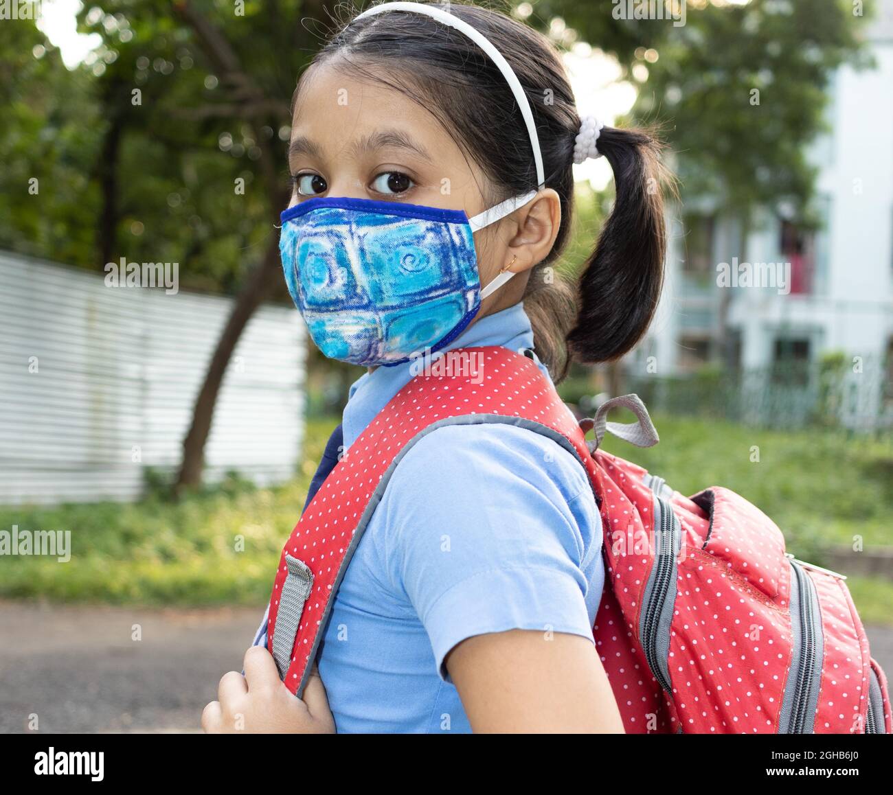 Close up of a happy Indian girl child student in blue school uniform with red bag and nose mask protection going to school Stock Photo