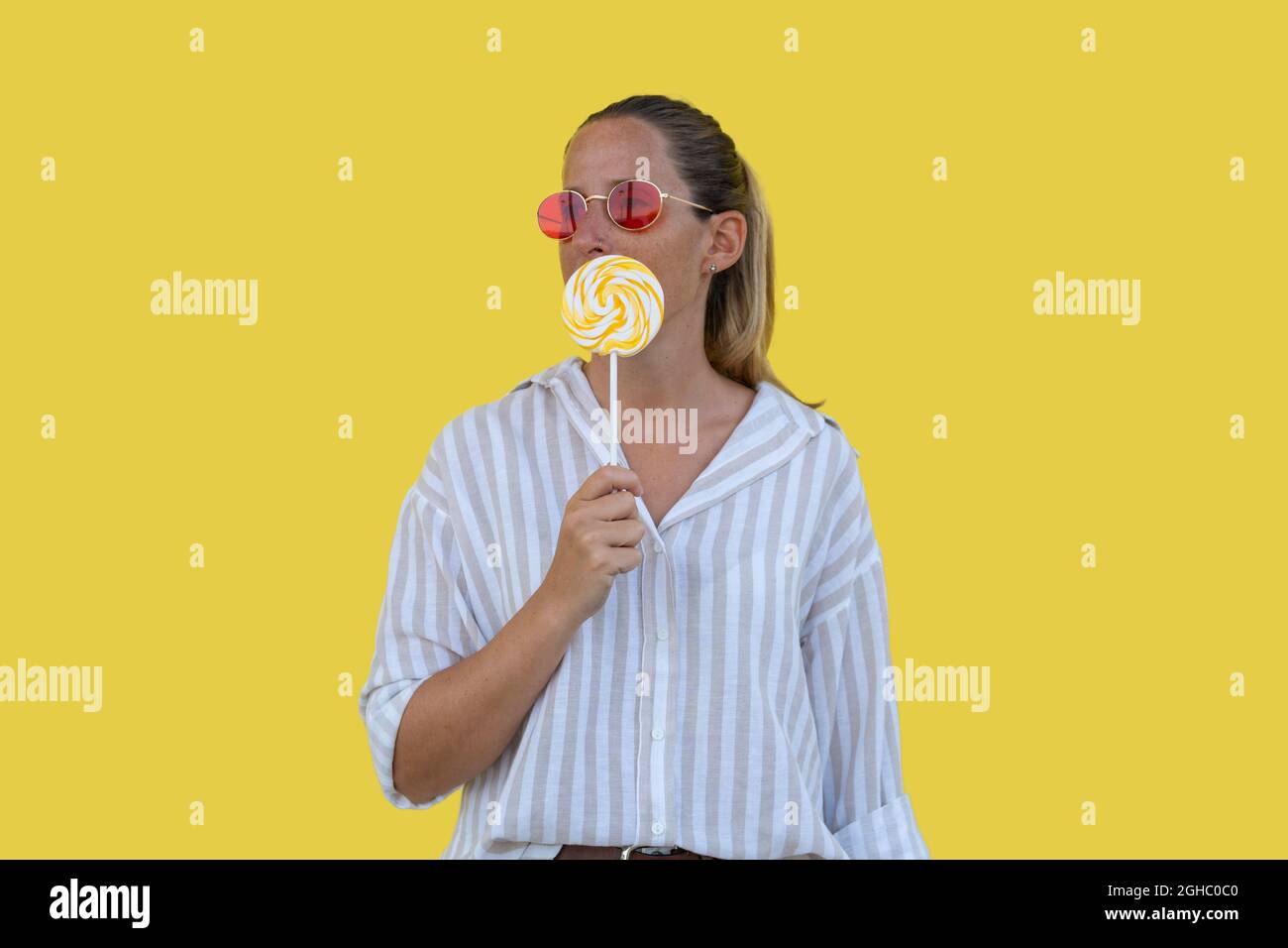 Female portrait in red sunglasses holding lollipop on yellow background Stock Photo