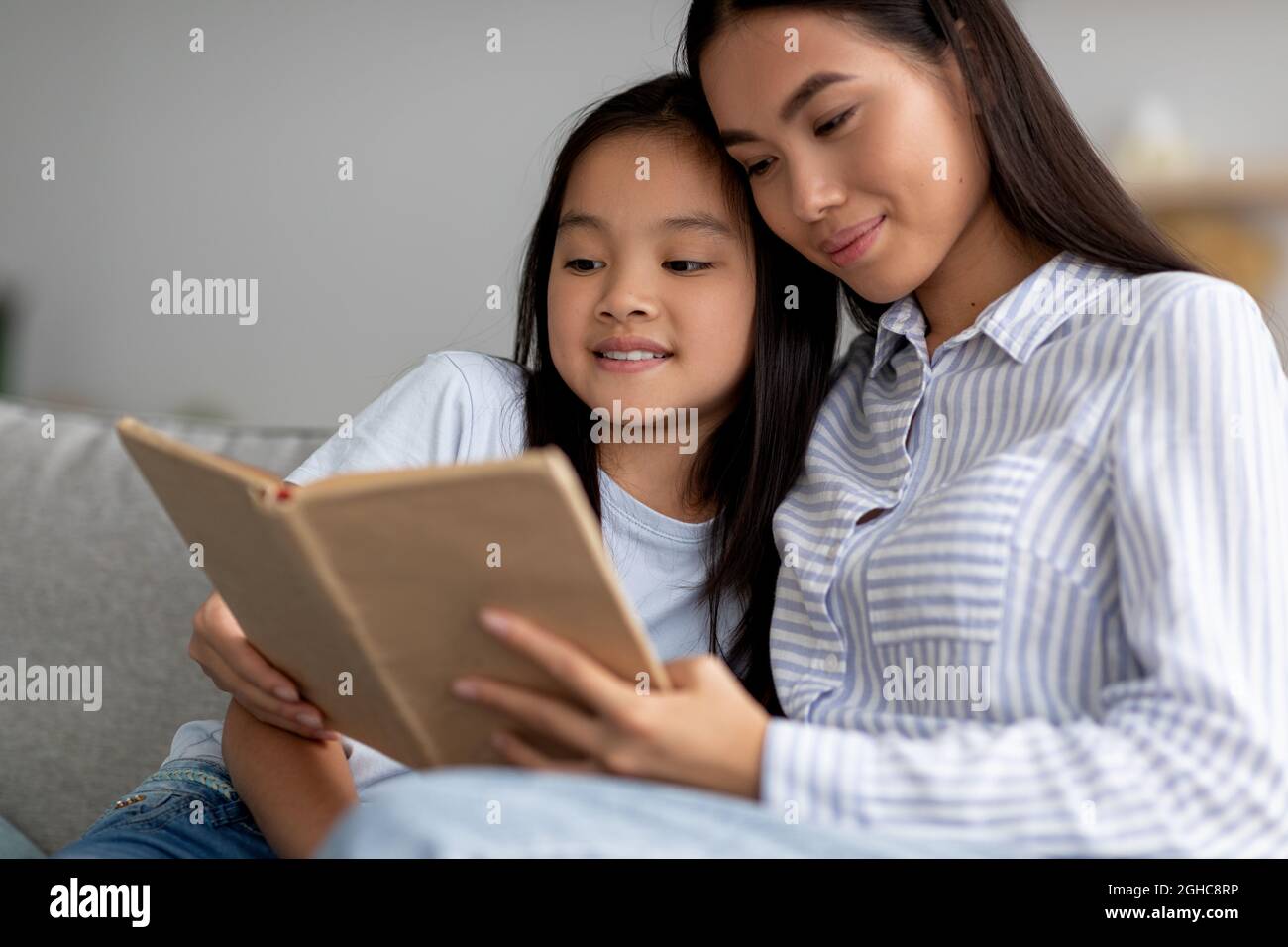 Happy asian girl reading book with her mother, sitting together on sofa and smiling, spending time at home interior Stock Photo