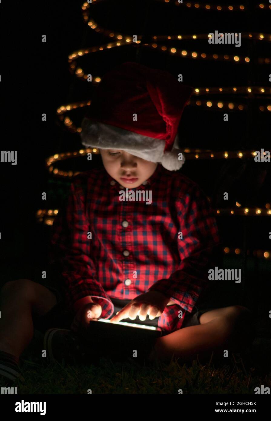 Brunette little boy wearing a red santa claus hat looking concentrated down at his tablet with the light on his face and christmas tree lights behind Stock Photo