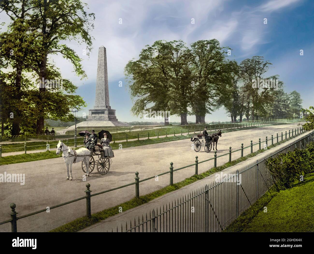 An early 20th century view of sightseers on a pony and trap passing the Wellington Monument, located in the Phoenix Park, Dublin City, Ireland. The 62 metres (203 ft) tall obelisk designed by Robert Smirke, commemorates the victories of the Duke of Wellington and is the largest obelisk in Europe. Stock Photo