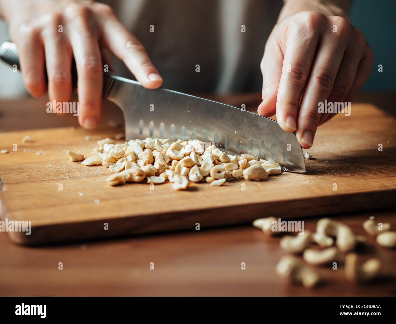 Female hands chopping cashew. Close up shot of process crushing raw unroasted cashew with large knife on wooden cutting board. Shallow DOF. Selective focus Stock Photo