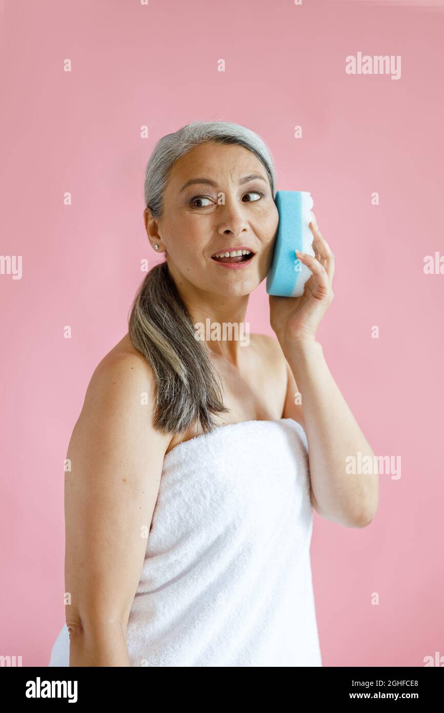 Beautiful hoary haired Asian woman uses bath sponge as mobile phone on pink background Stock Photo