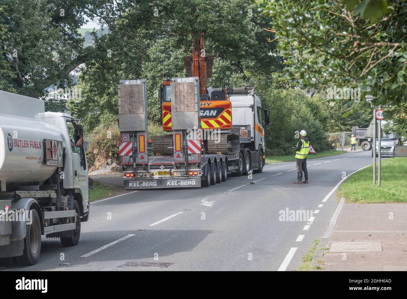 Low loader with construction site excavator being unloaded at a civil engineering works entrance and causing a road blockage and traffic tailback. Stock Photo