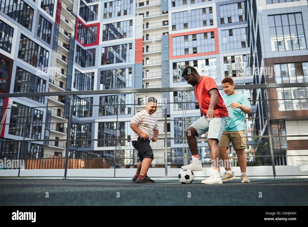 Cheerful young black man playing soccer with boys against modern flat block building Stock Photo