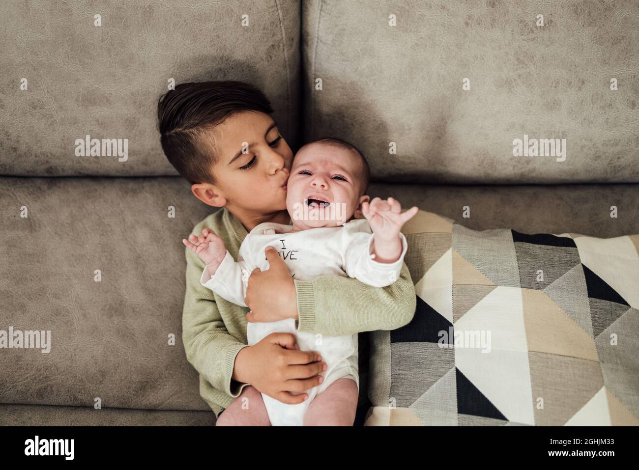Young boy kissing his baby sister on the cheek while lying together on the sofa at home. Stock Photo