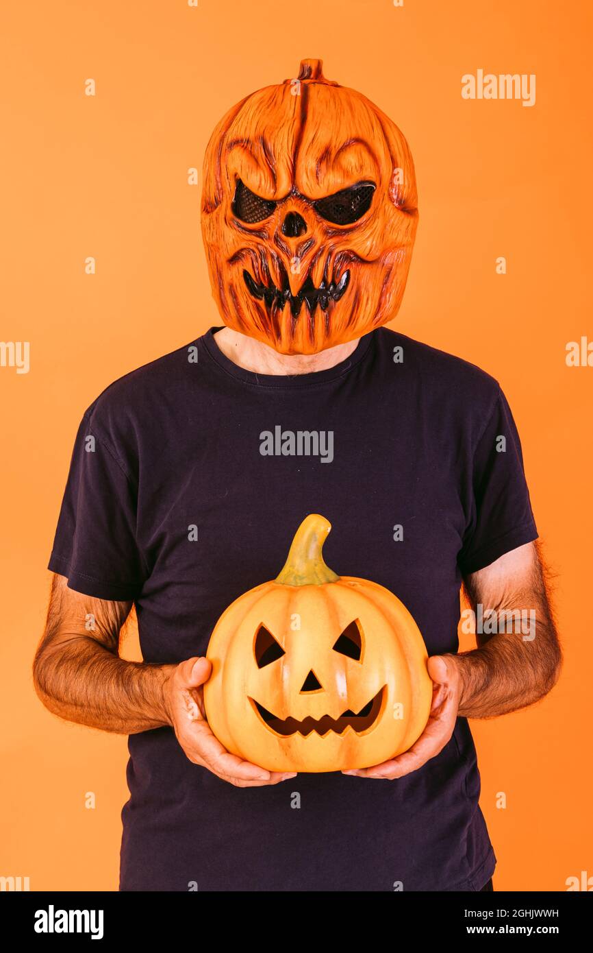 Man wearing scary pumpkin latex mask with blue t-shirt, holding 'Jack-o-lantern' pumpkin on orange background. Halloween and days of the dead concept. Stock Photo