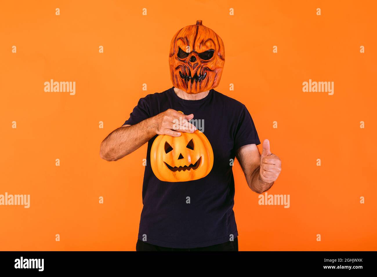 Man wearing scary pumpkin latex mask with blue t-shirt, holds a 'Jack-o-lantern' pumpkin and thumbs up, on orange background. Halloween and days of th Stock Photo