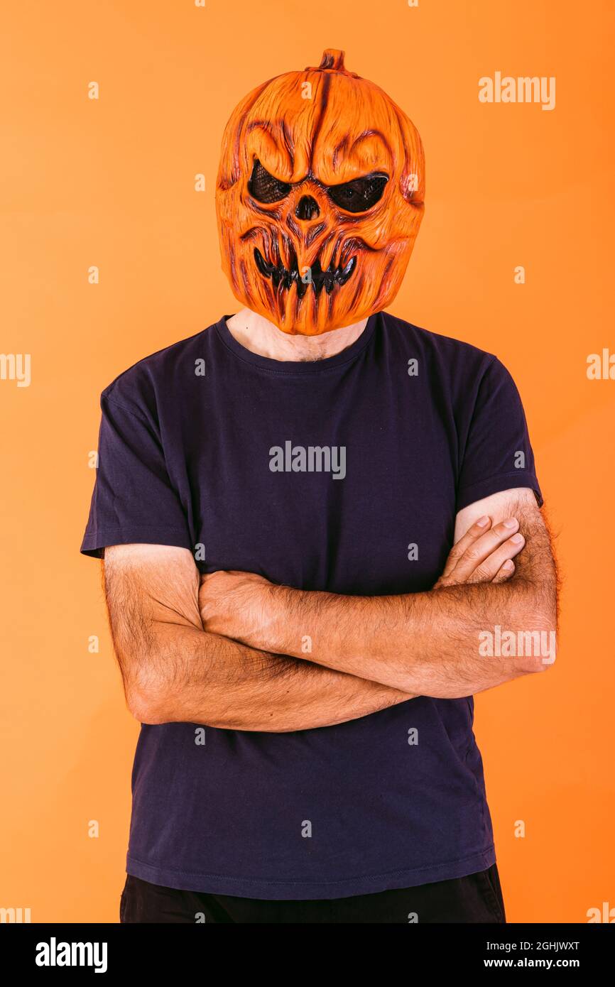 Man wearing scary pumpkin latex mask with blue t-shirt with crossed arms, on orange background. Halloween and days of the dead concept. Stock Photo