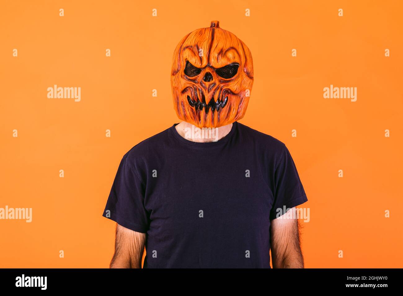 Man wearing scary pumpkin latex mask with blue t-shirt on orange background. Halloween and days of the dead concept. Stock Photo