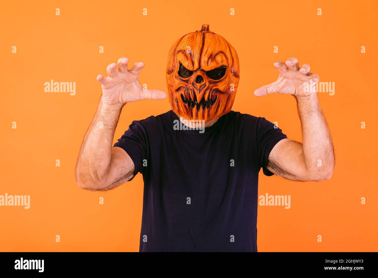 Man wearing scary pumpkin latex mask with blue t-shirt scares with his hands, on orange background. Halloween and days of the dead concept. Stock Photo