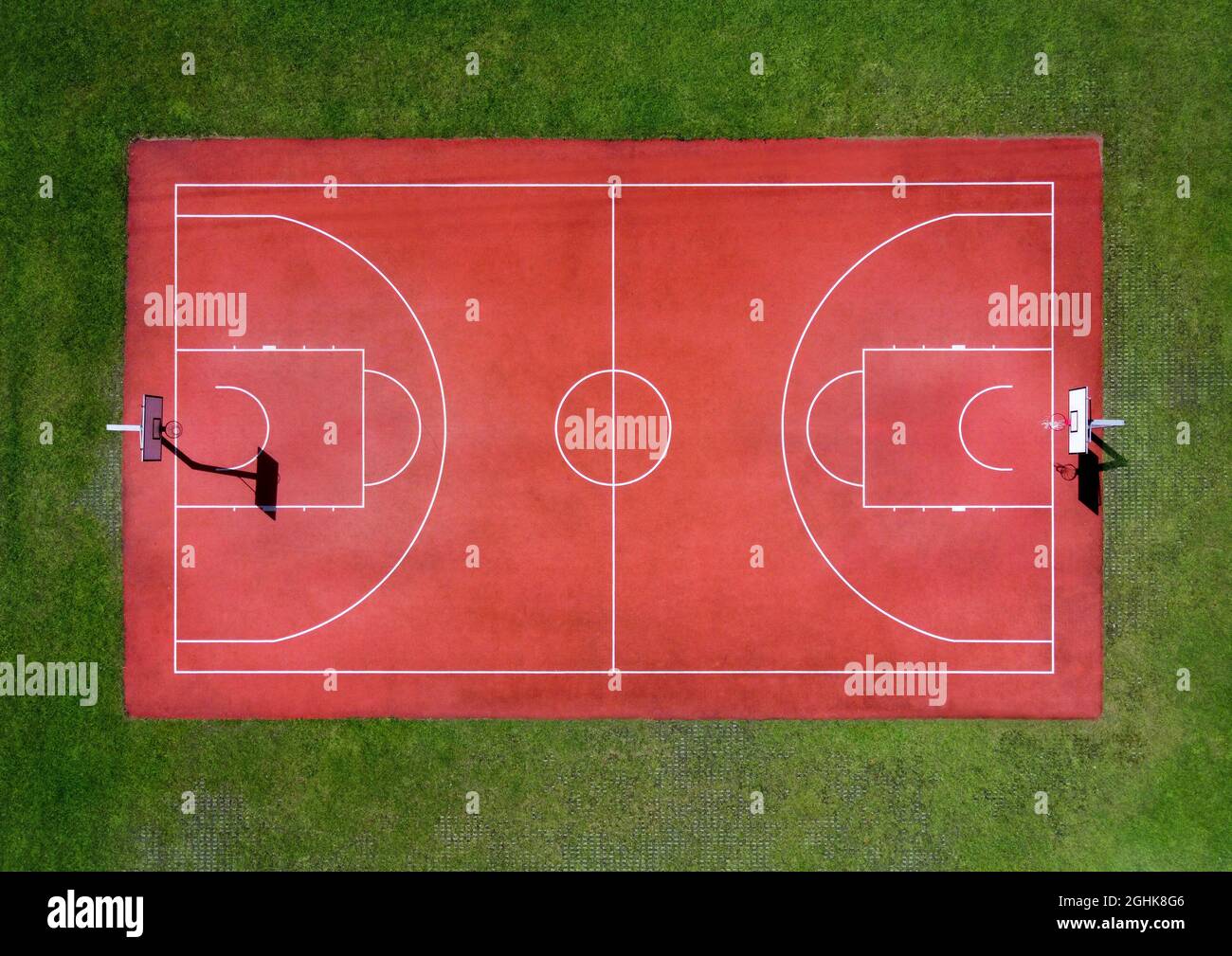 From above aerial view of empty red basketball sports field with white markings and hoops surrounded by green grass Stock Photo