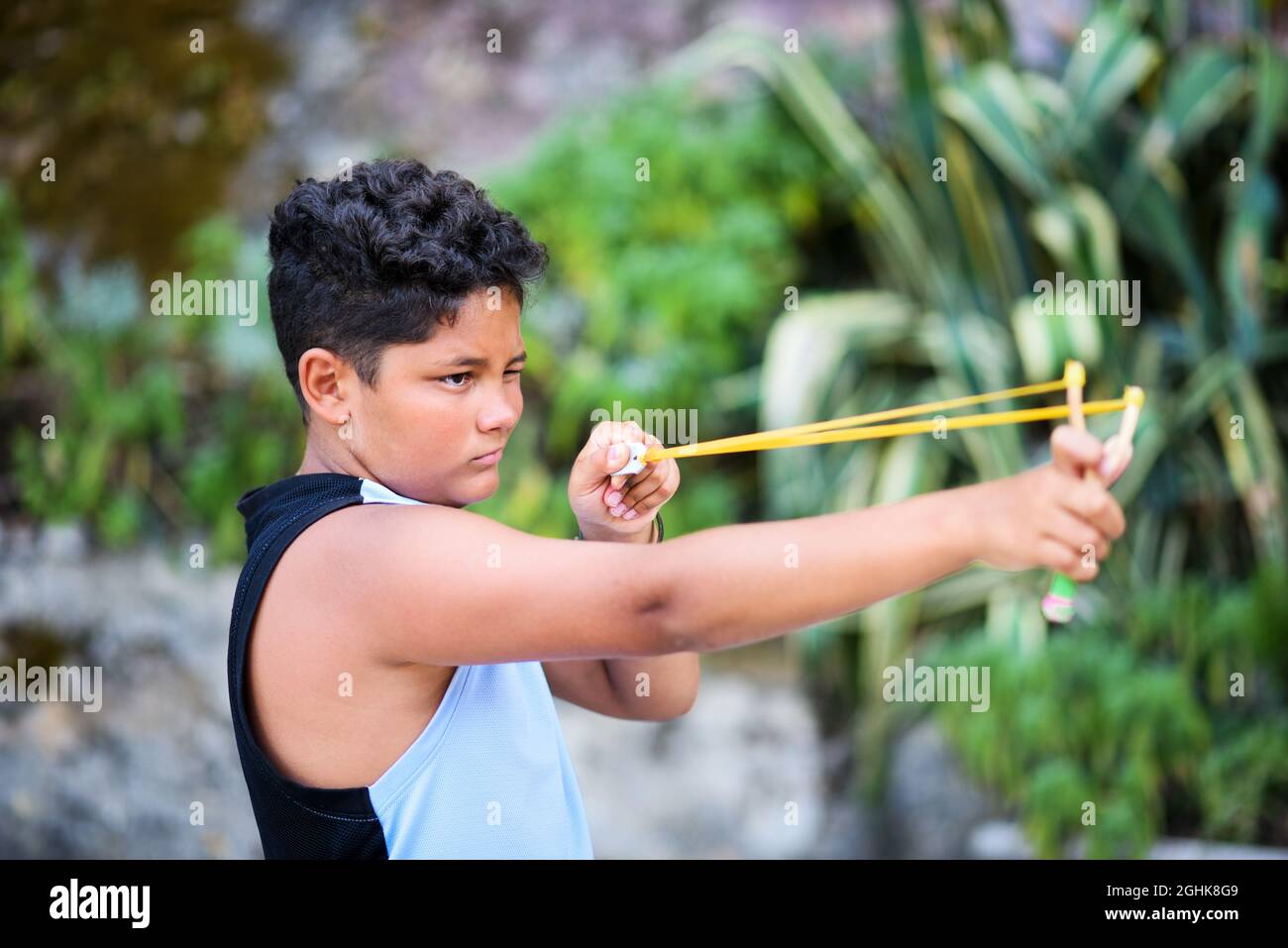 Boy with curly hair closing eyes and aiming while playing with sling on blurred background of garden on summer weekend day Stock Photo