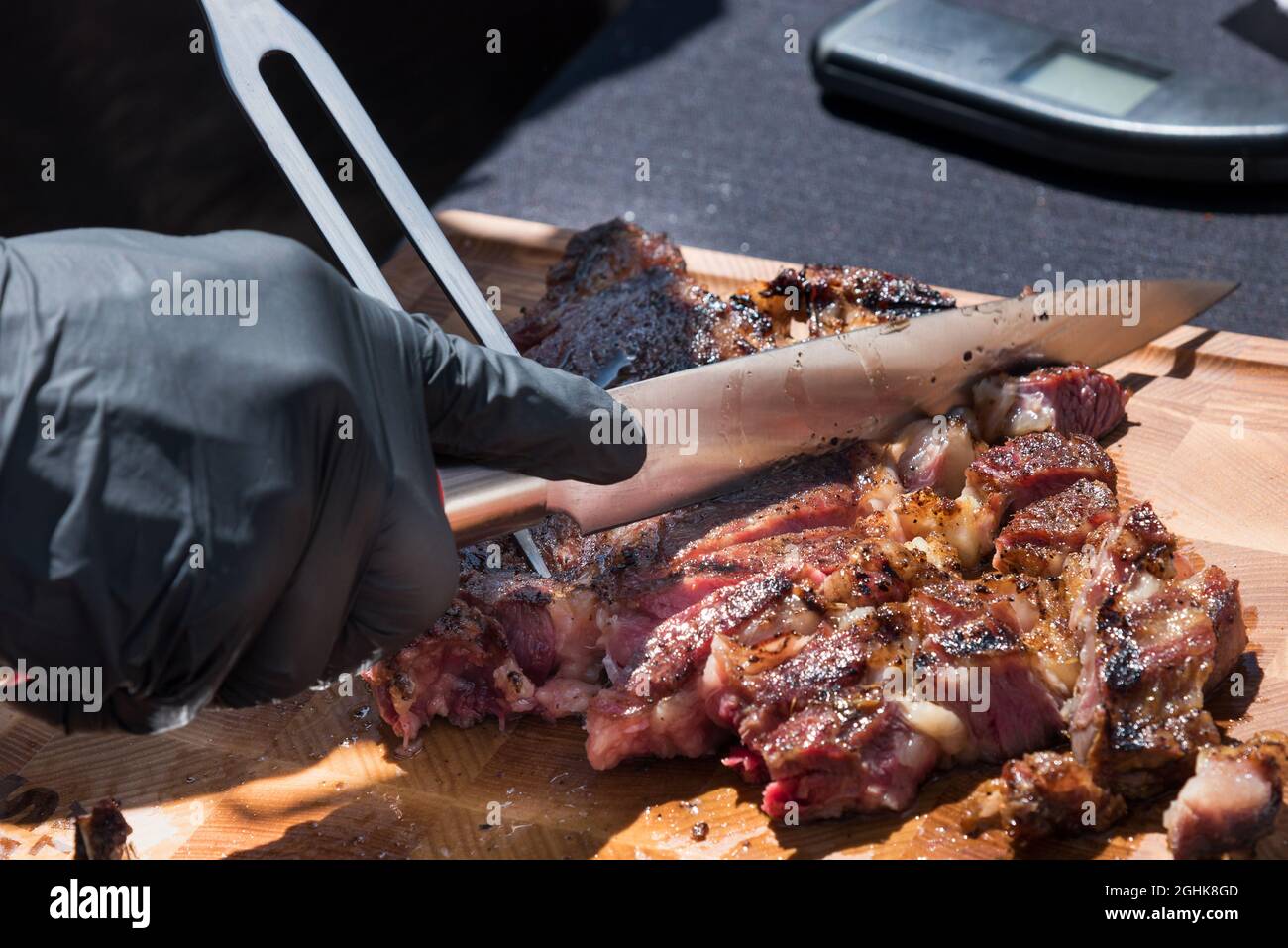 Gloved hand of a chef carving a portion of grilled prime rib eye meat from the BBQ on a wooden chopping board in a close up on the food Stock Photo