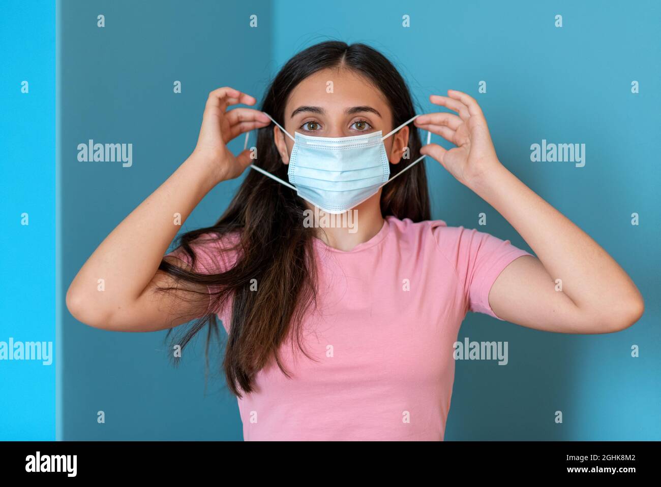 Teen female putting on protective medical mask for coronavirus prevention against blue background Stock Photo