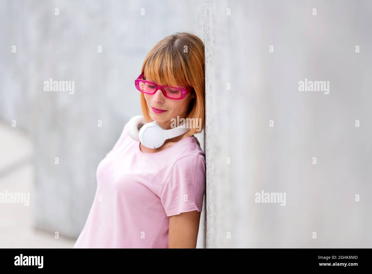 Trendy woman in pink glasses standing relaxing against a wall looking down with a quiet smile of contentment, with lateral copyspace Stock Photo