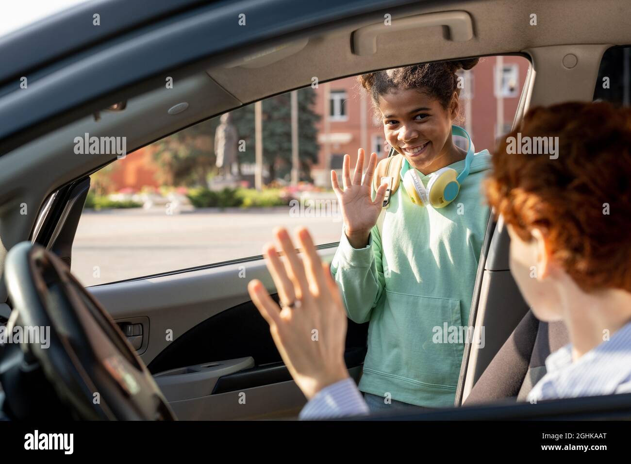 Smiling schoolgirl waving hand to her mom while getting out of car before going to school Stock Photo