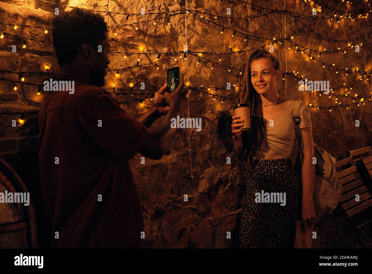 Happy young female with drink posing for her African boyfriend with smartphone in the evening Stock Photo