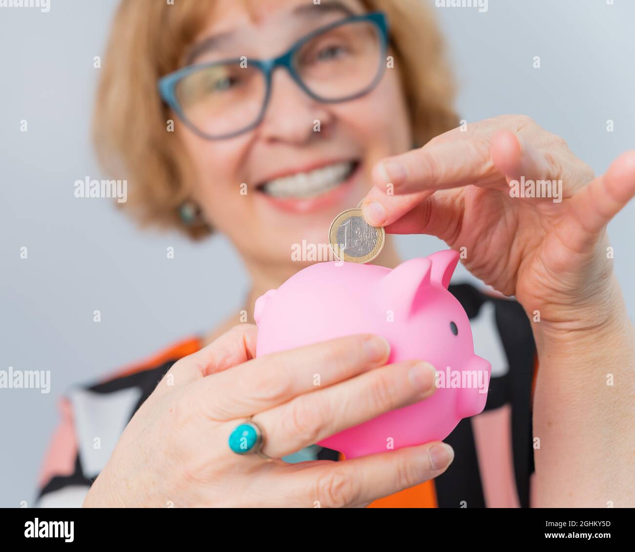 An elderly woman with glasses puts a coin in a piggy bank on a white ...