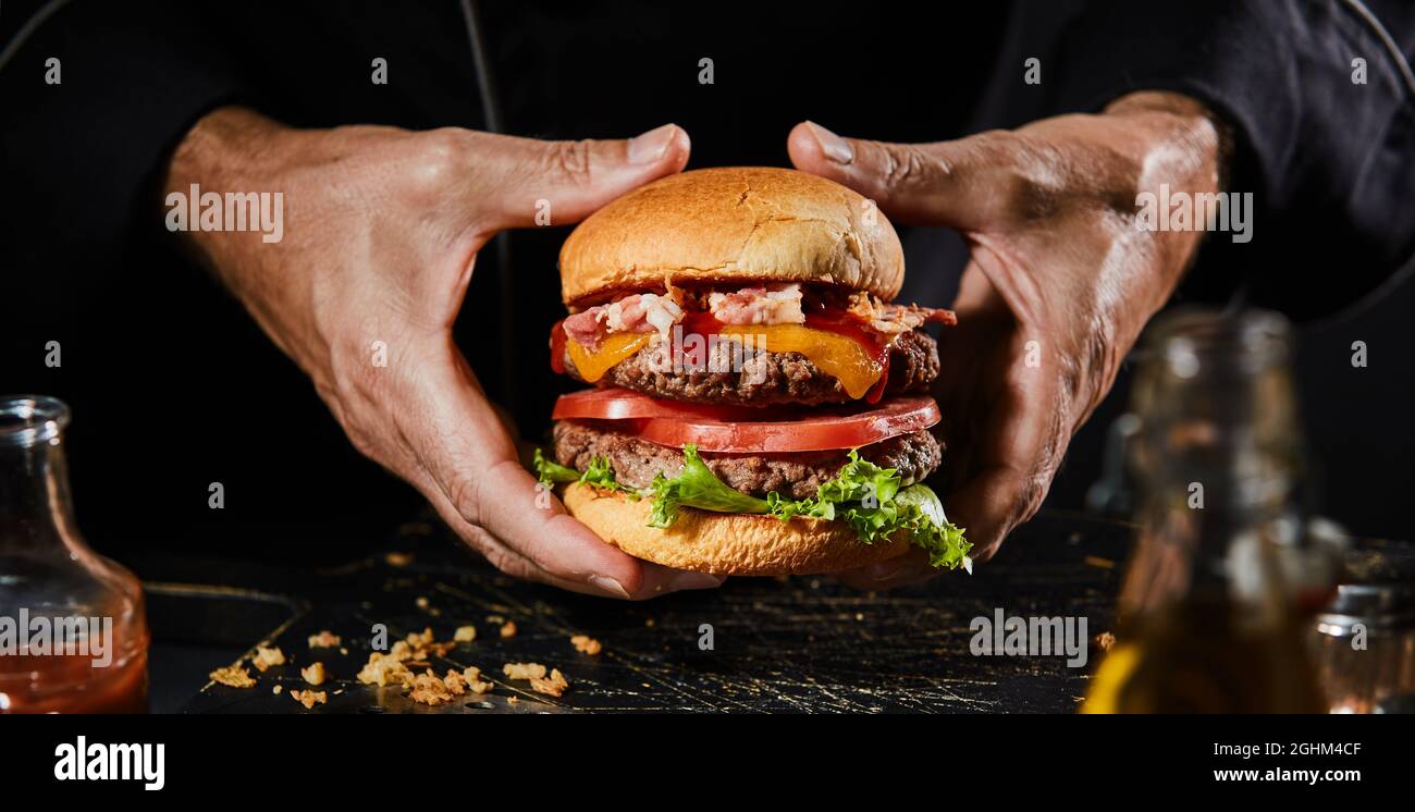Panorama banner with hands of a man holding a double beef burger, whopper or Big Boy with bacon, salad and cheese trimmings on a toasted bun for menu Stock Photo