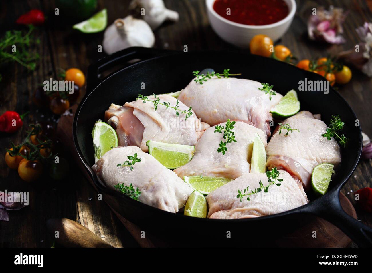 Raw uncooked chicken thigh meat in a cast iron frying pan with fresh ingredients of limes, spicy sauce, lemon thyme, peppers, tomatoes and garlic. Stock Photo