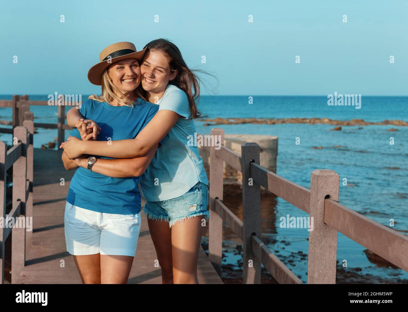 Daughter embracing Mother and standing on wooden sidewalk by the sea at sunset. A middle-aged woman and teenage girl standing side by side and wearing Stock Photo