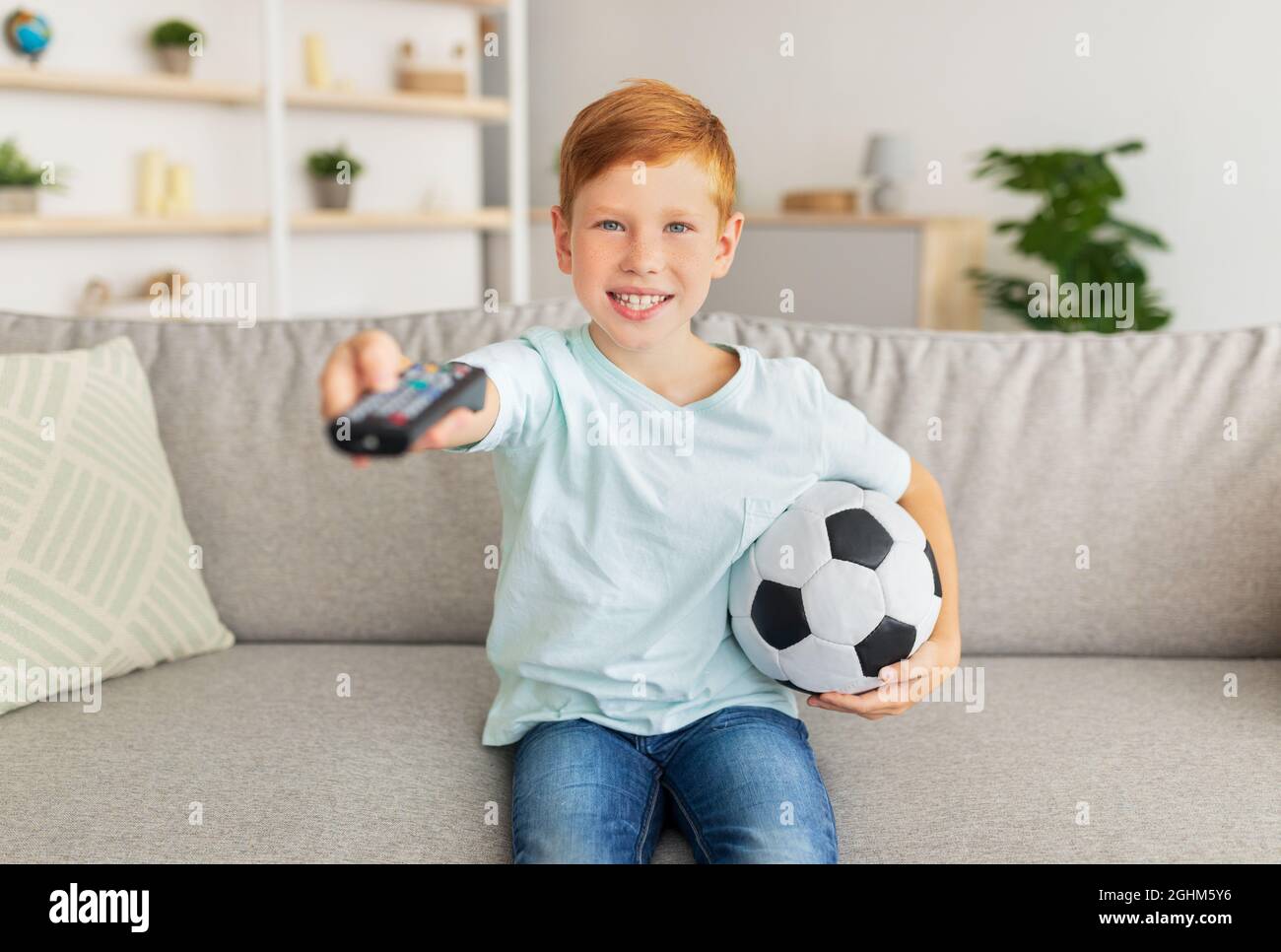 Happy redhead boy with ball watching football game on TV Stock Photo