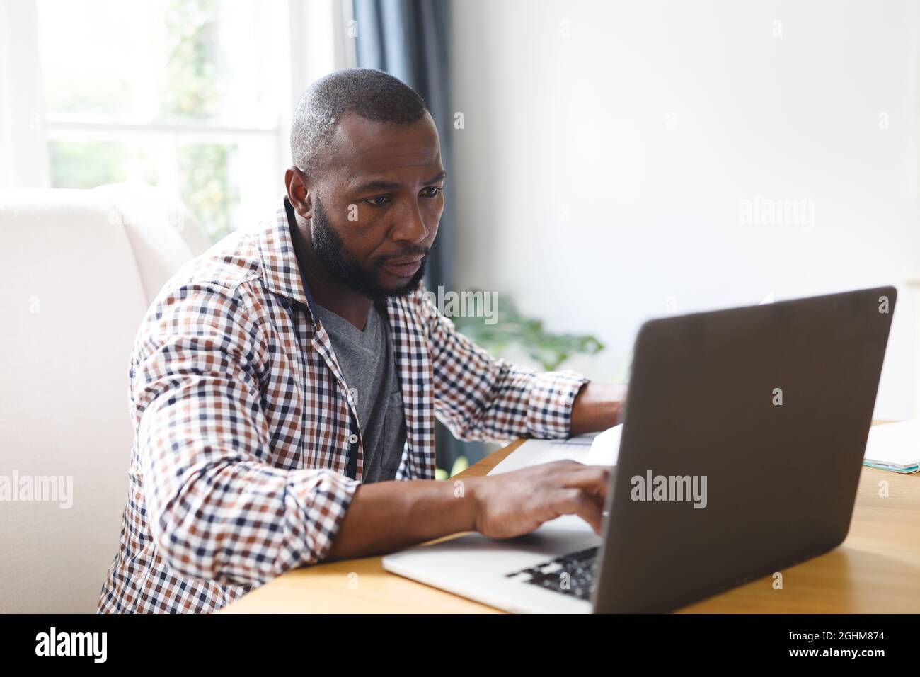 African american man sitting at table in dining room, working remotely using laptop Stock Photo