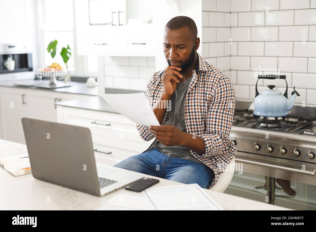 Serious african american man sitting in kitchen working looking at paperwork and using laptop Stock Photo