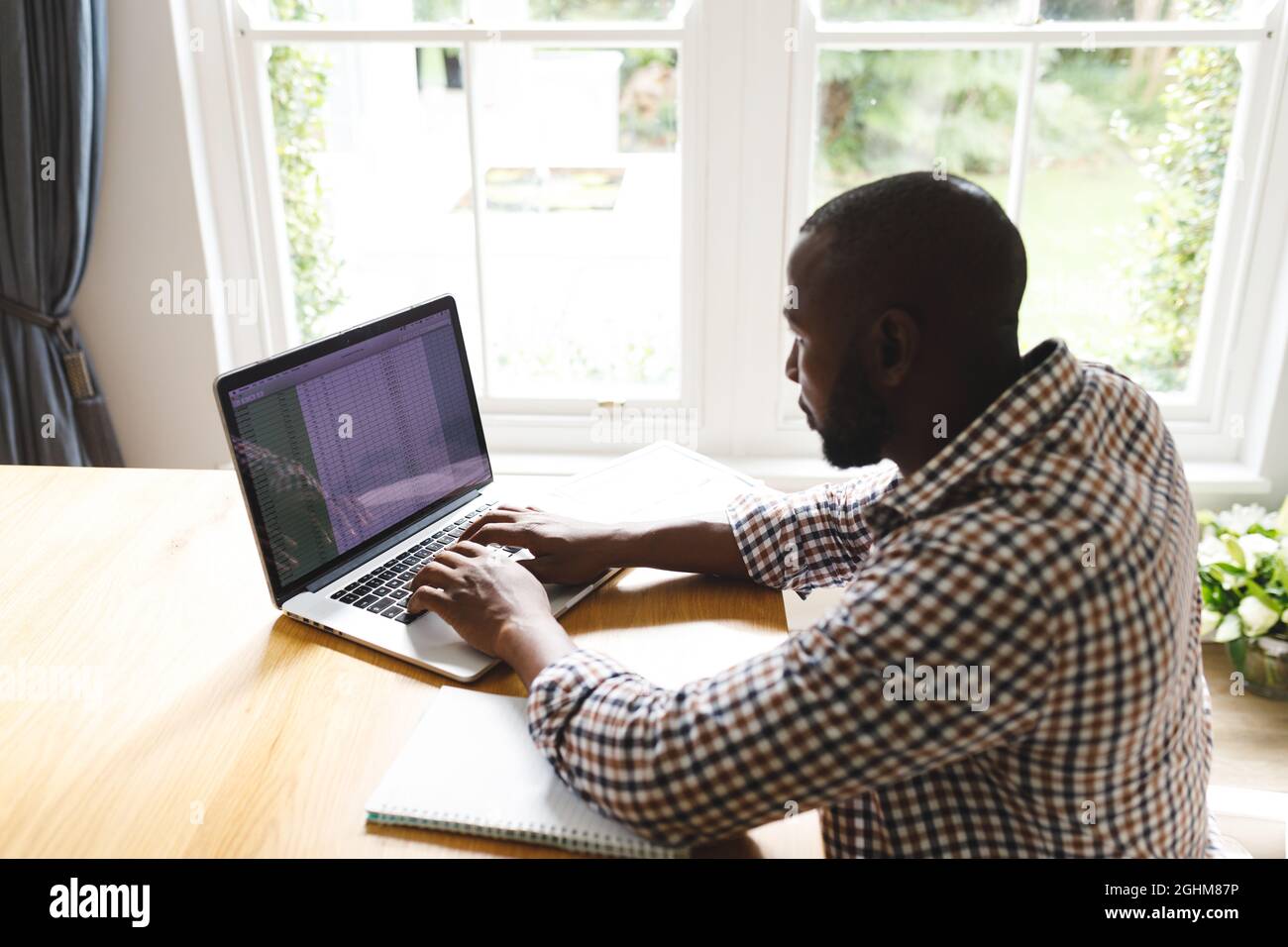 African american man sitting at table in dining room, working remotely using laptop Stock Photo