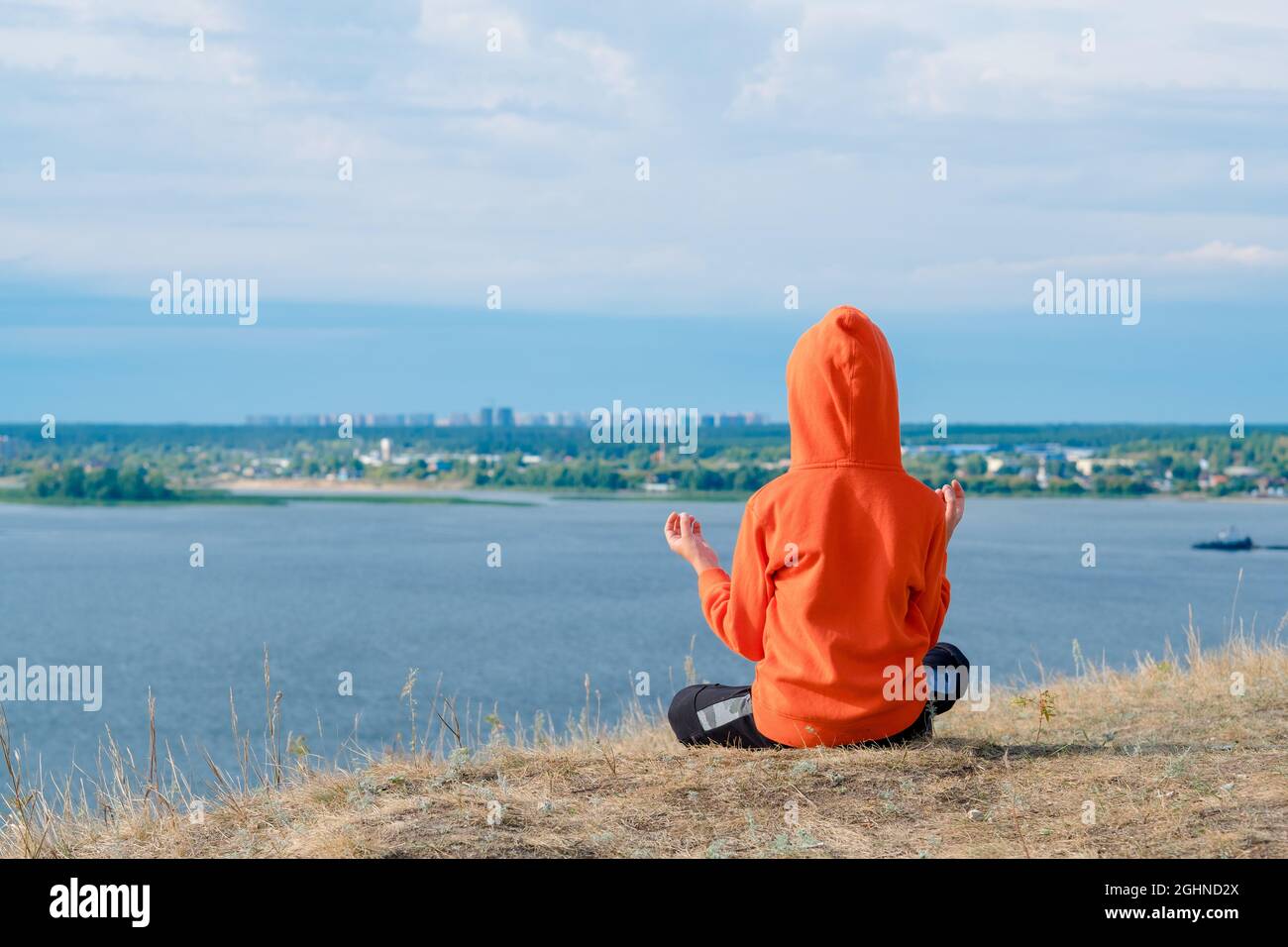 boy meditates on the hillside against the background of the sea. Beautiful seascape with a meditating boy from the back. Mental health from childhood. Stock Photo